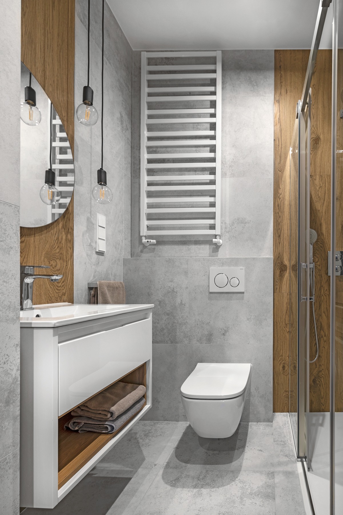 Nowoczesna łazienka z szarymi płytkami, drewnianymi akcentami i białymi meblami, oświetlona wiszącymi lampami z żarówkami Edisona.