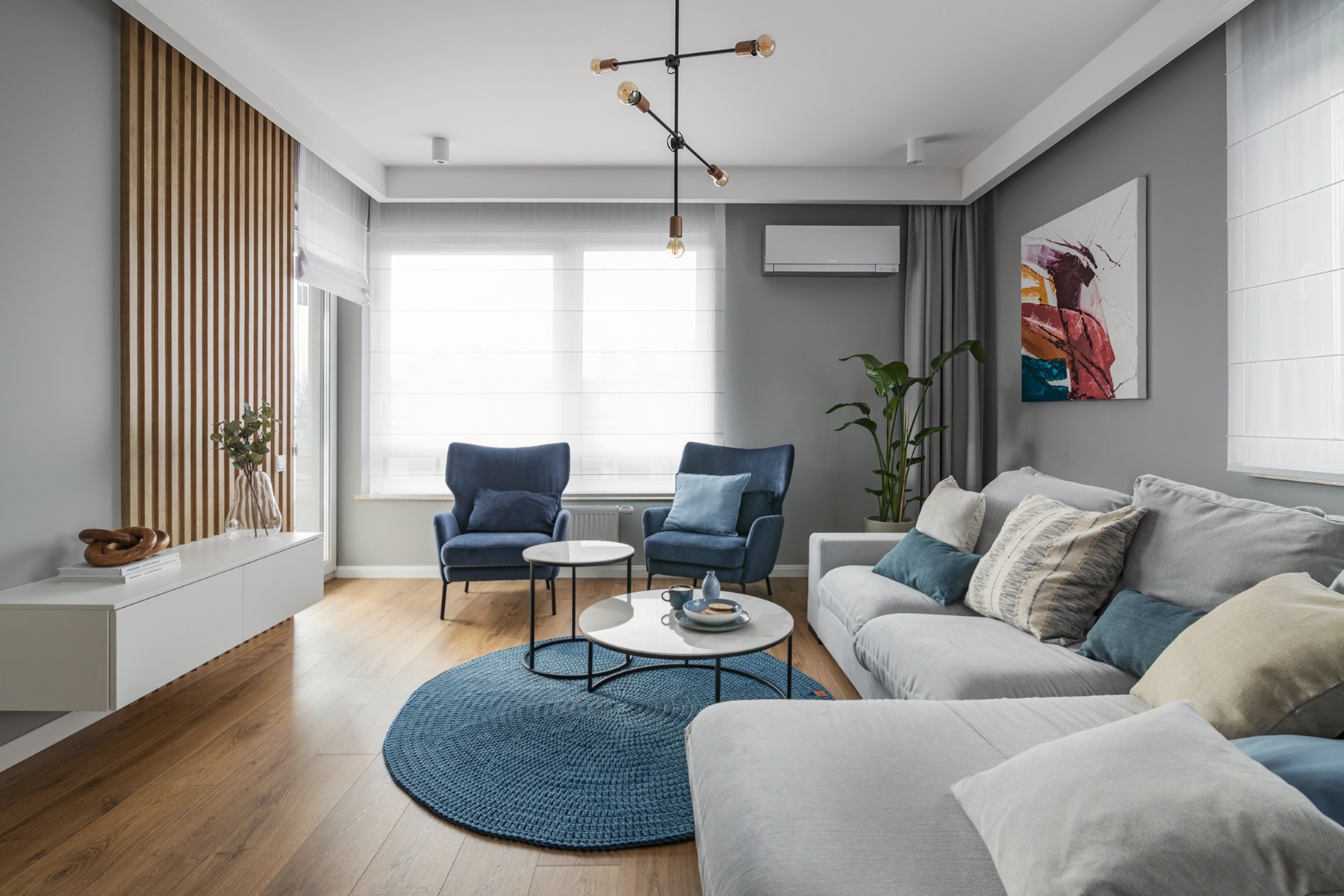 Jasne wnętrze salonu z drewnianą podłogą, szarą sofą narożną z poduszkami, dwoma granatowymi fotelami, okrągłym niebieskim dywanem, stolikami kawowymi z marmurowym blatem, drewnianą ścianką...