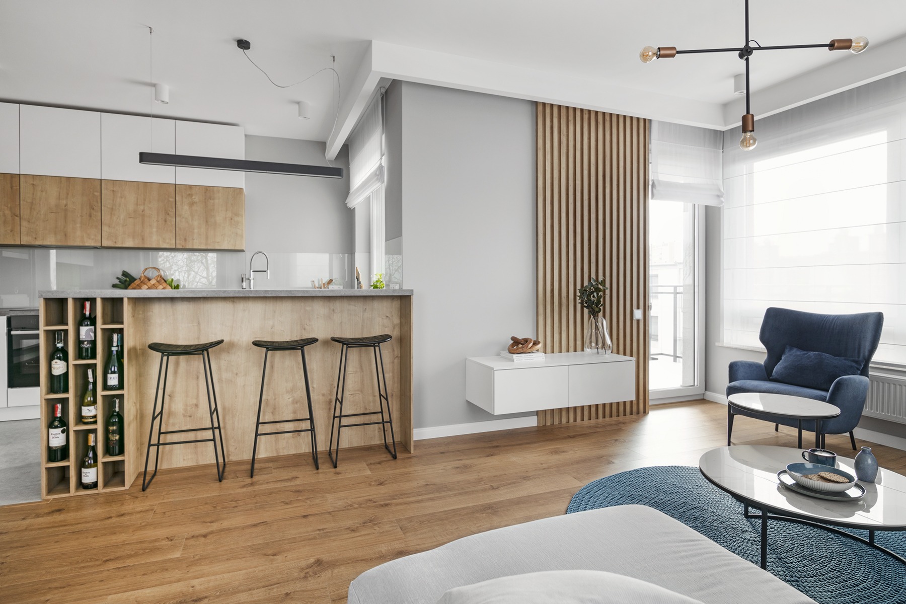 Jasne wnętrze z drewnianą podłogą i elementami, biało-drewniana zabudowa kuchenna z wyspą i barkiem, trzy czarne hokery, regał na wino, drewniana ażurowa ściana, biała szafka RTV, granatowy fotel...