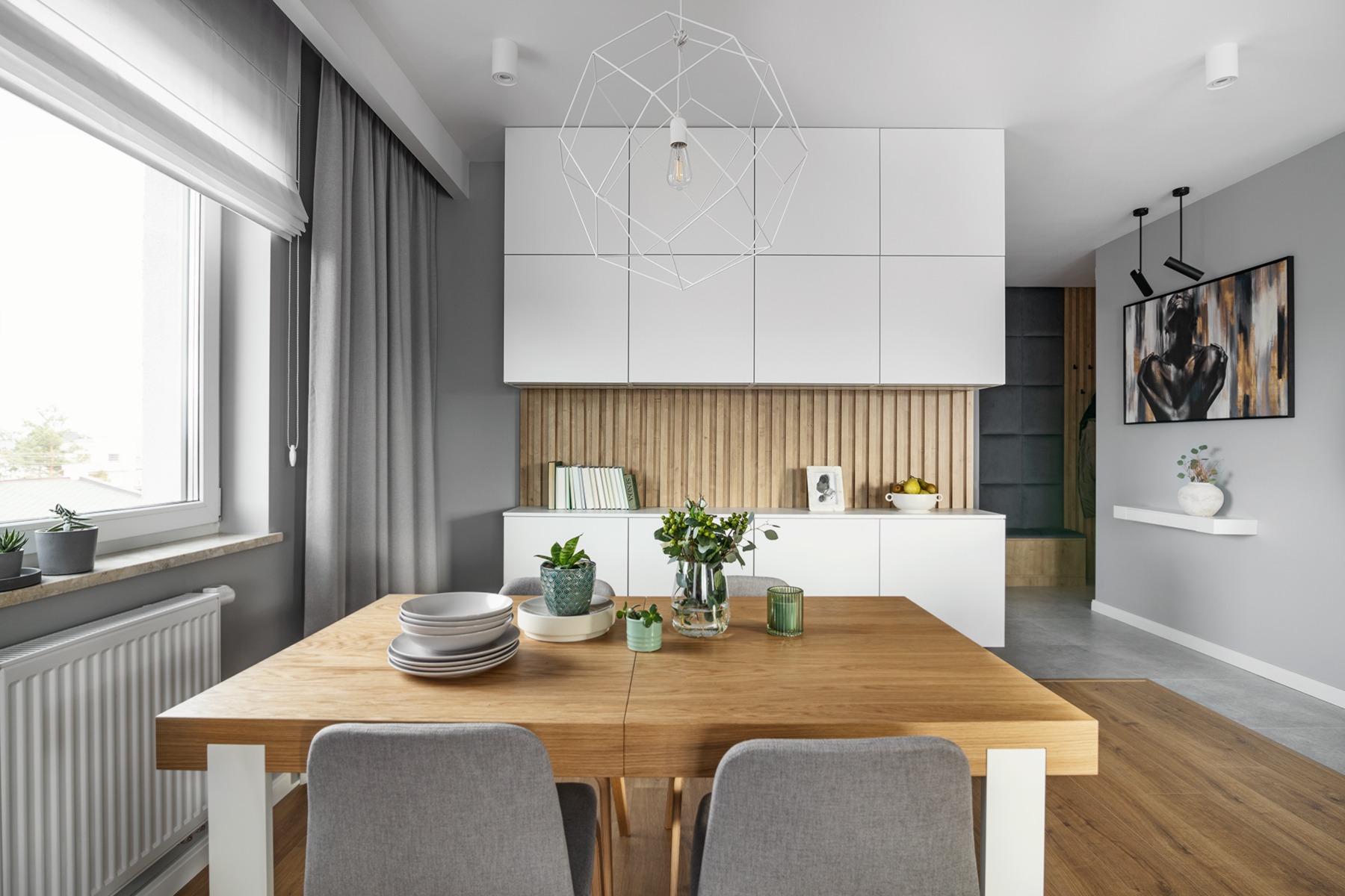 Jasne wnętrze jadalni z drewnianym stołem, szarymi krzesłami i geometryczną lampą wiszącą, białe szafki w tle z drewnianą okładziną ścienną.