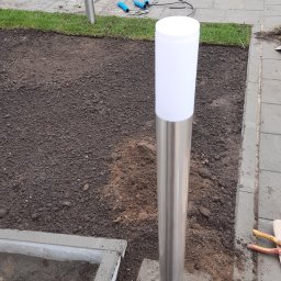 VK-LAD ELECTRICS SP. Z O.O. - Nowo zamontowana, nowoczesna lampa ogrodowa z metalową obudową i białym kloszem, umieszczona na betonowej podstawie w ziemi. Widoczne narzędzia i kable sugerują trwające prace wykończeniowe.