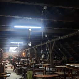 VK-LAD ELECTRICS SP. Z O.O. - Strych z drewnianą konstrukcją dachu, oświetlony szeregiem długich lamp jarzeniowych i wiszących żarówek, wypełniony starymi stołami i krzesłami.