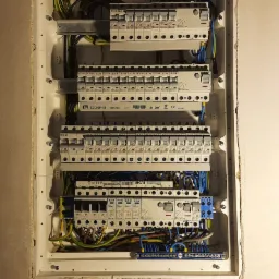 Wewnętrzna skrzynka rozdzielcza z zainstalowanymi bezpiecznikami i podłączonymi przewodami elektrycznymi, widoczna puszka instalacyjna w ścianie pod skrzynką.