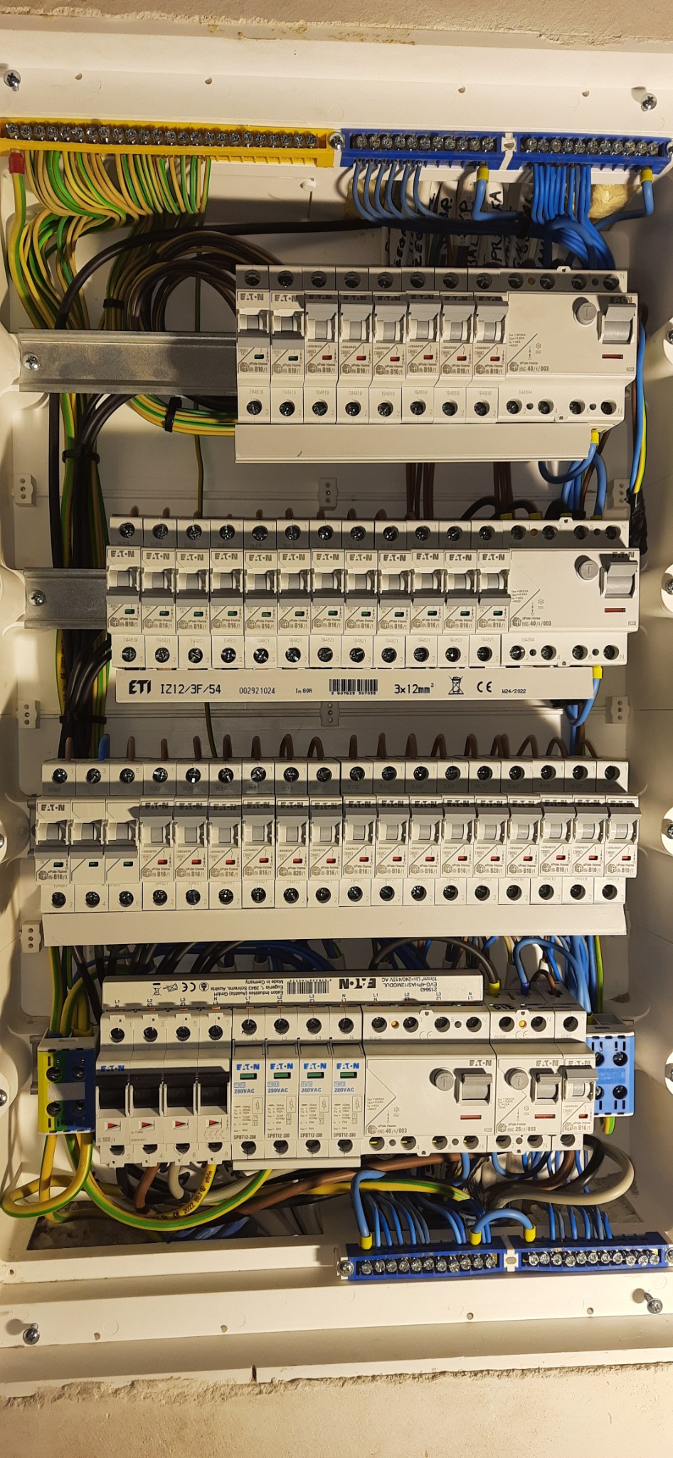 Rozdzielnica elektryczna z bezpiecznikami, przewodami w kolorach: żółto-zielonym, niebieskim i brązowym, widoczne elementy montażowe i oznaczenia producentów Eaton i ETI
