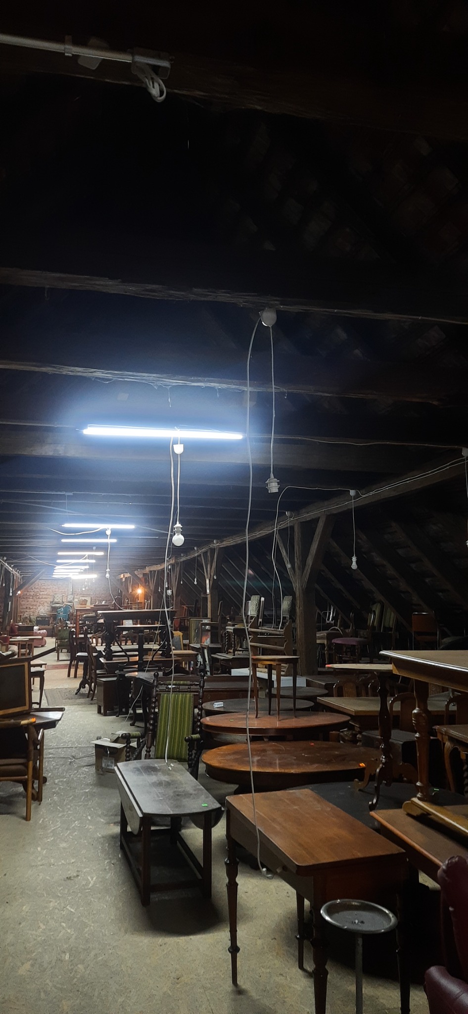 Strych z drewnianą konstrukcją dachu, oświetlony szeregiem długich lamp jarzeniowych i wiszących żarówek, wypełniony starymi stołami i krzesłami.