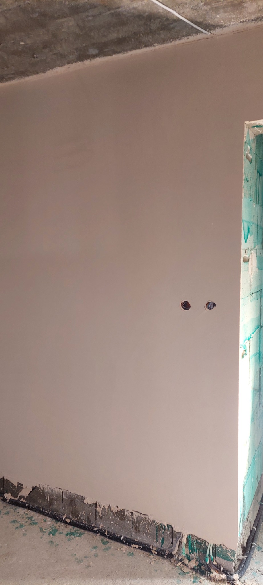 Świeżo otynkowana ściana wewnętrzna z widocznymi otworami na gniazdka elektryczne, dolna krawędź ściany z resztkami zaprawy i przewodami, surowy betonowy sufit, narożnik zabezpieczony folią budowlaną.