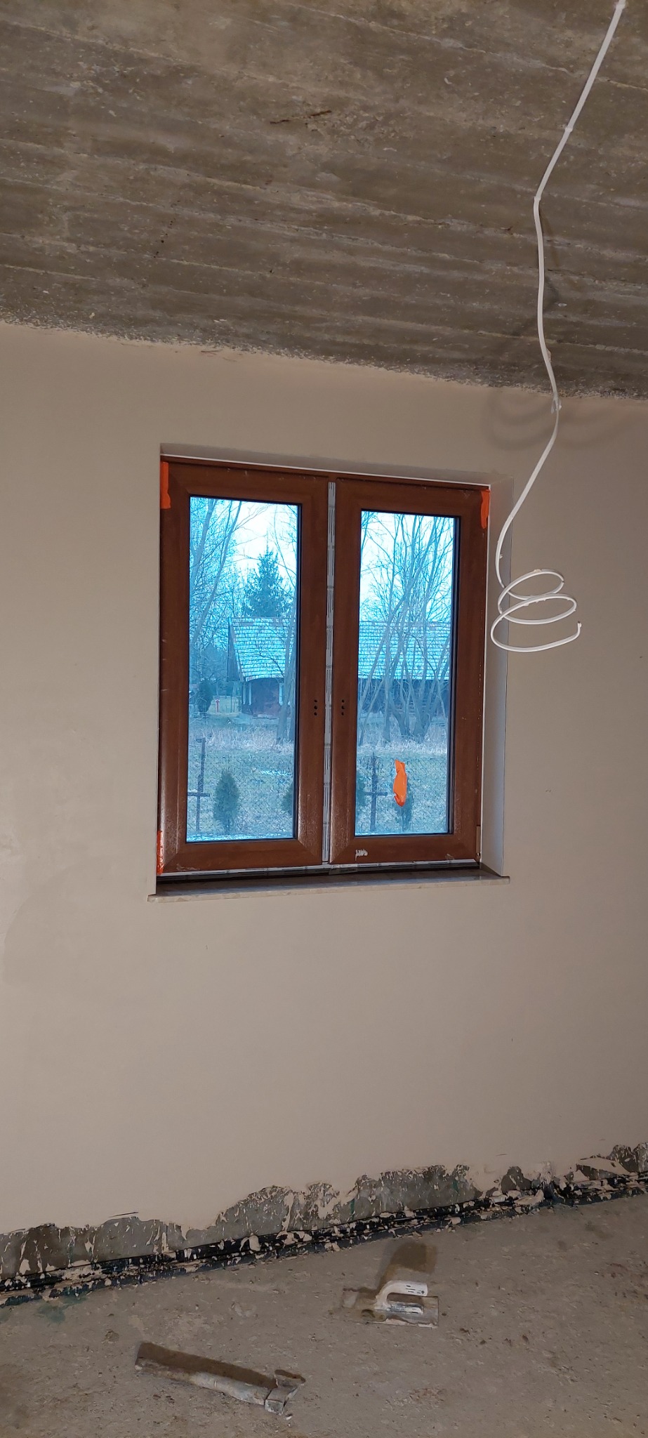 Pokój w trakcie wykończenia: nowo wstawione okno z brązowymi ramami, ściany po gładzi, widoczne niedoskonałości przy podłodze i wystająca instalacja elektryczna z sufitu.