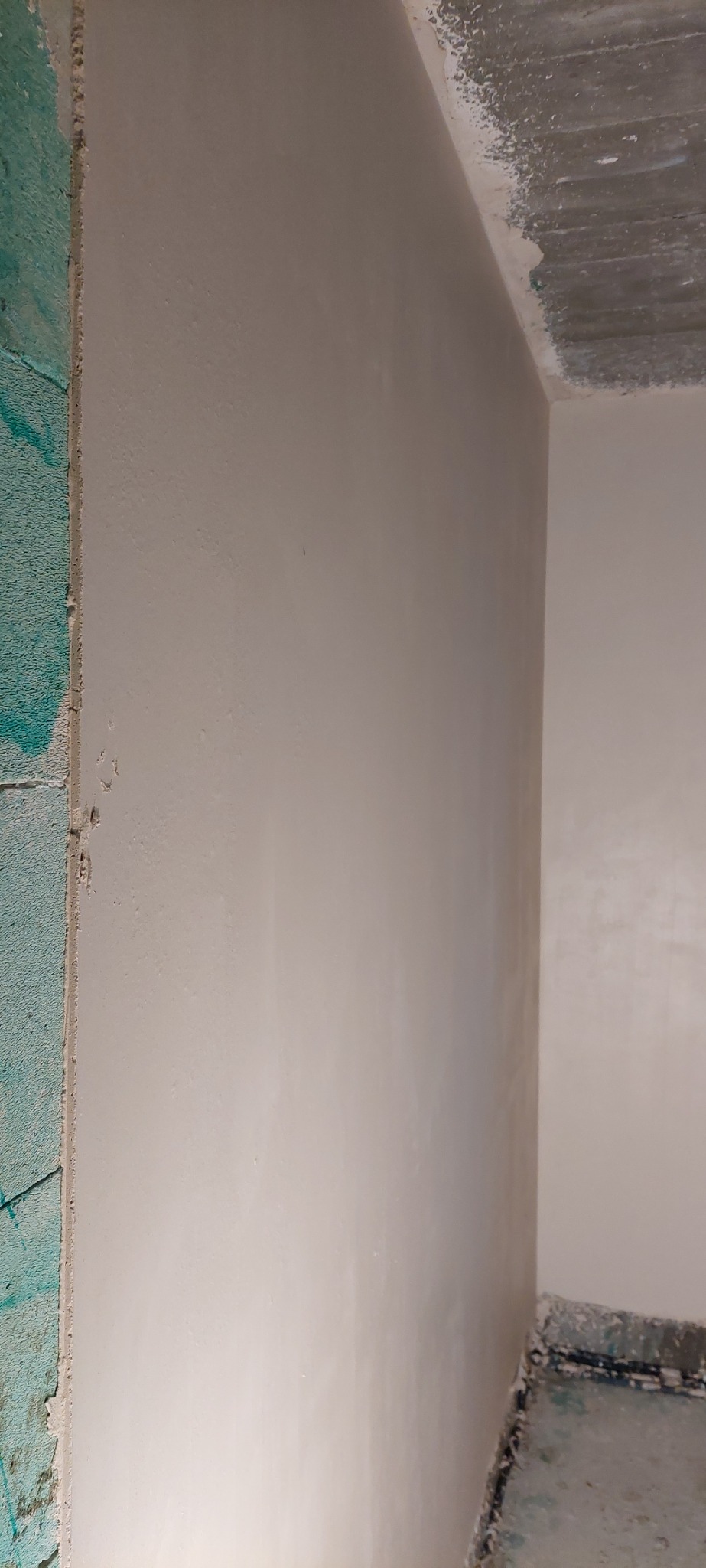 Świeżo otynkowana ściana w rogu pomieszczenia, z widocznym surowym betonowym sufitem i fragmentem nieotynkowanej ściany z zielonymi blokami.