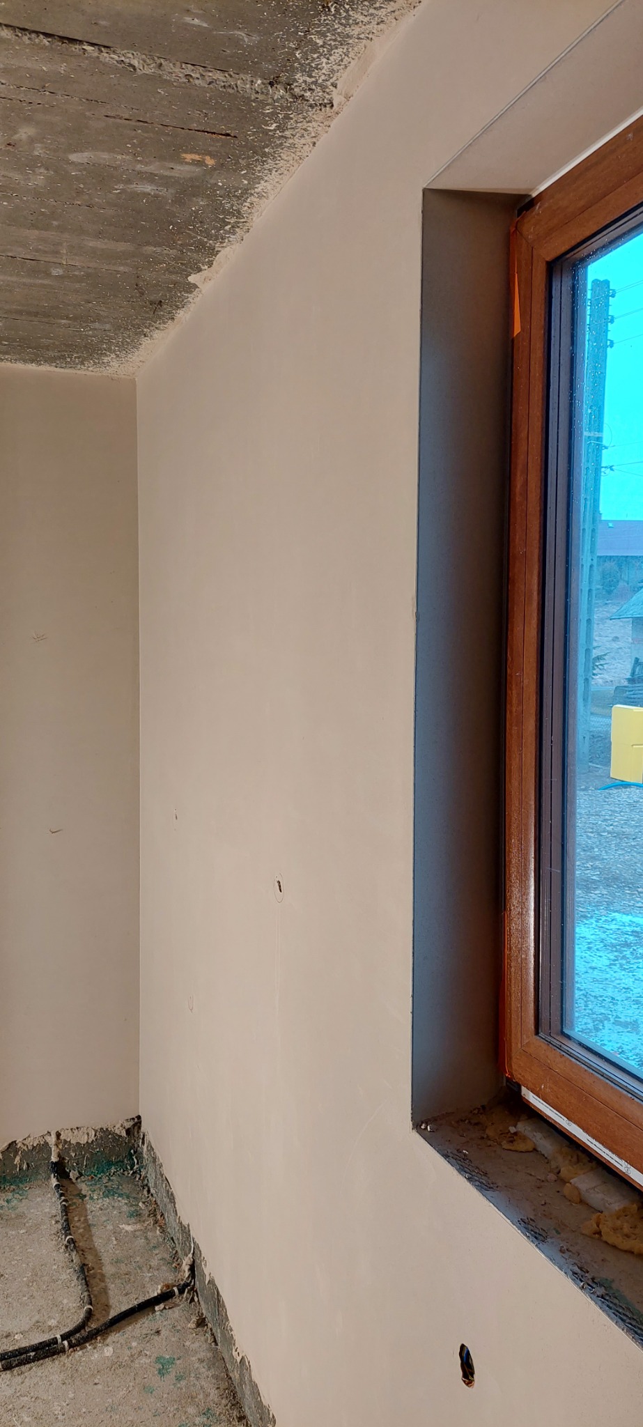 Świeżo otynkowane ściany w pomieszczeniu z drewnianym oknem, widoczne nierówności sufitu i instalacje przy podłodze w trakcie prac wykończeniowych.