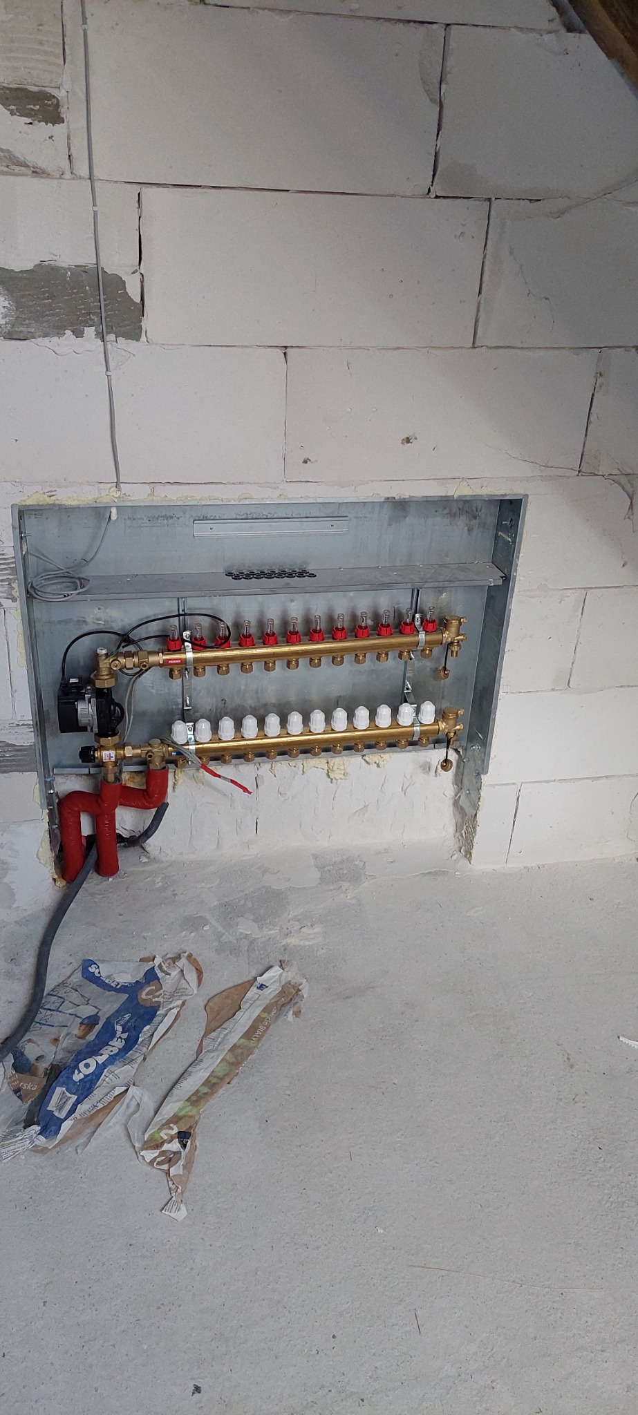 Rozdzielacz ogrzewania podłogowego zamontowany w ścianie z bloczków, widoczne rury w izolacji, elementy instalacji i worki po materiałach budowlanych na posadzce.
