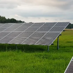 Panel słoneczny na metalowej konstrukcji, otoczony zieloną trawą, z widocznymi balotami siana w tle i pochmurnym niebem.