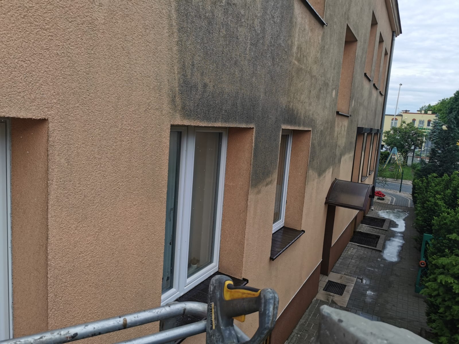 Fragment elewacji budynku wielorodzinnego w trakcie czyszczenia, widoczna wyraźna różnica między umytą, jasną powierzchnią a zabrudzoną, ciemniejszą częścią. W tle widoczne okna, chodnik i zieleń.