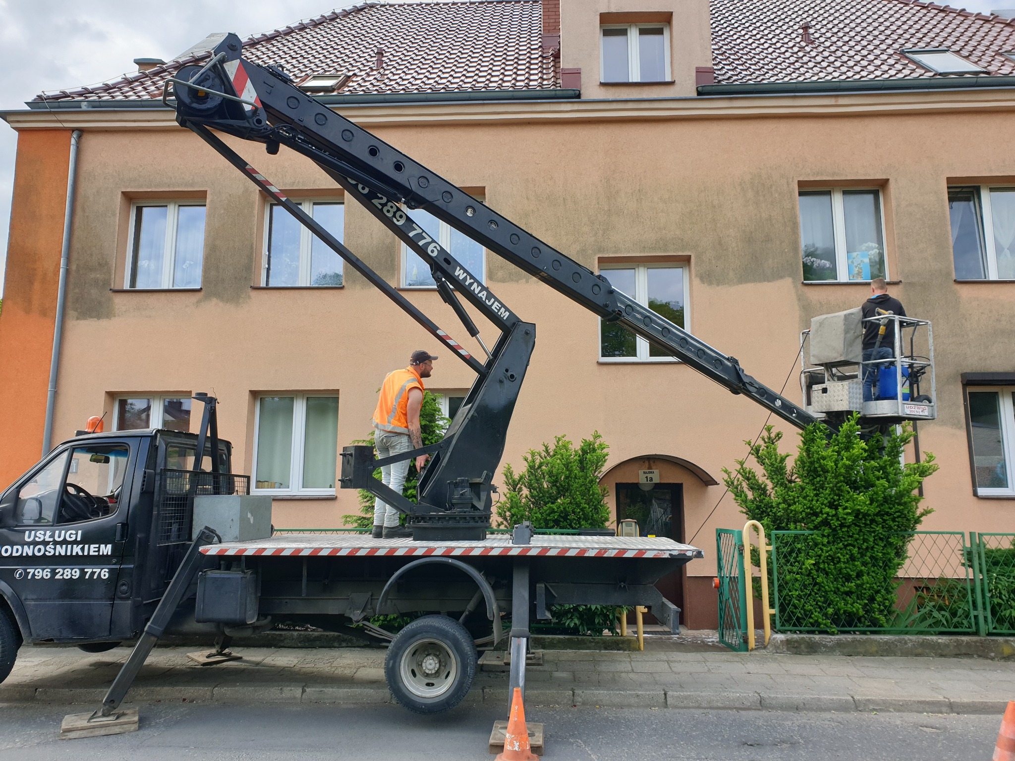 Pracownicy na podnośniku koszowym czyszczą elewację budynku mieszkalnego, widoczne różnice w kolorze elewacji przed i po czyszczeniu.