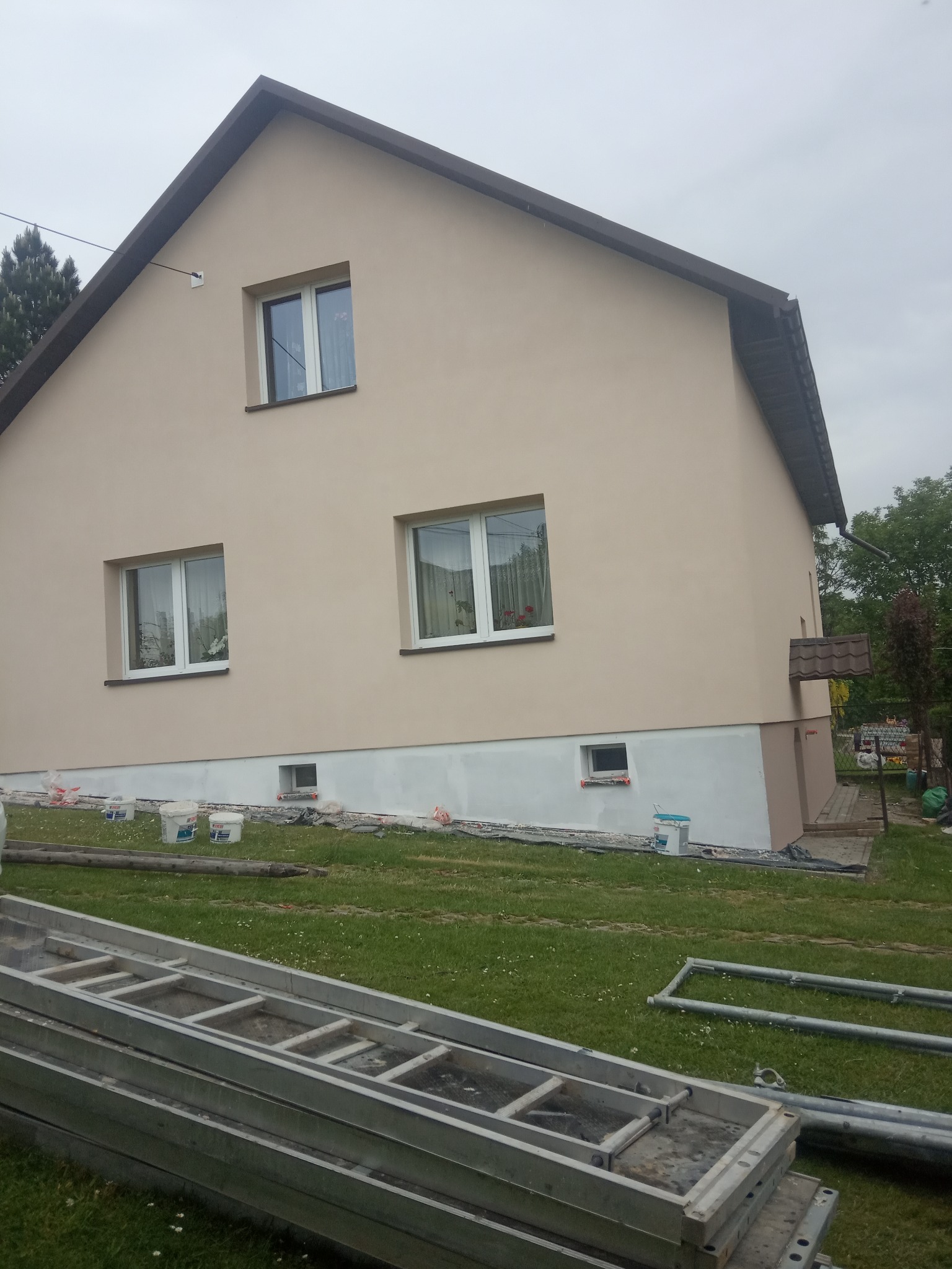 Dom jednorodzinny z beżową elewacją, widoczne ślady prac przy cokole, leżące drabiny i wiaderka z farbą na trawniku.