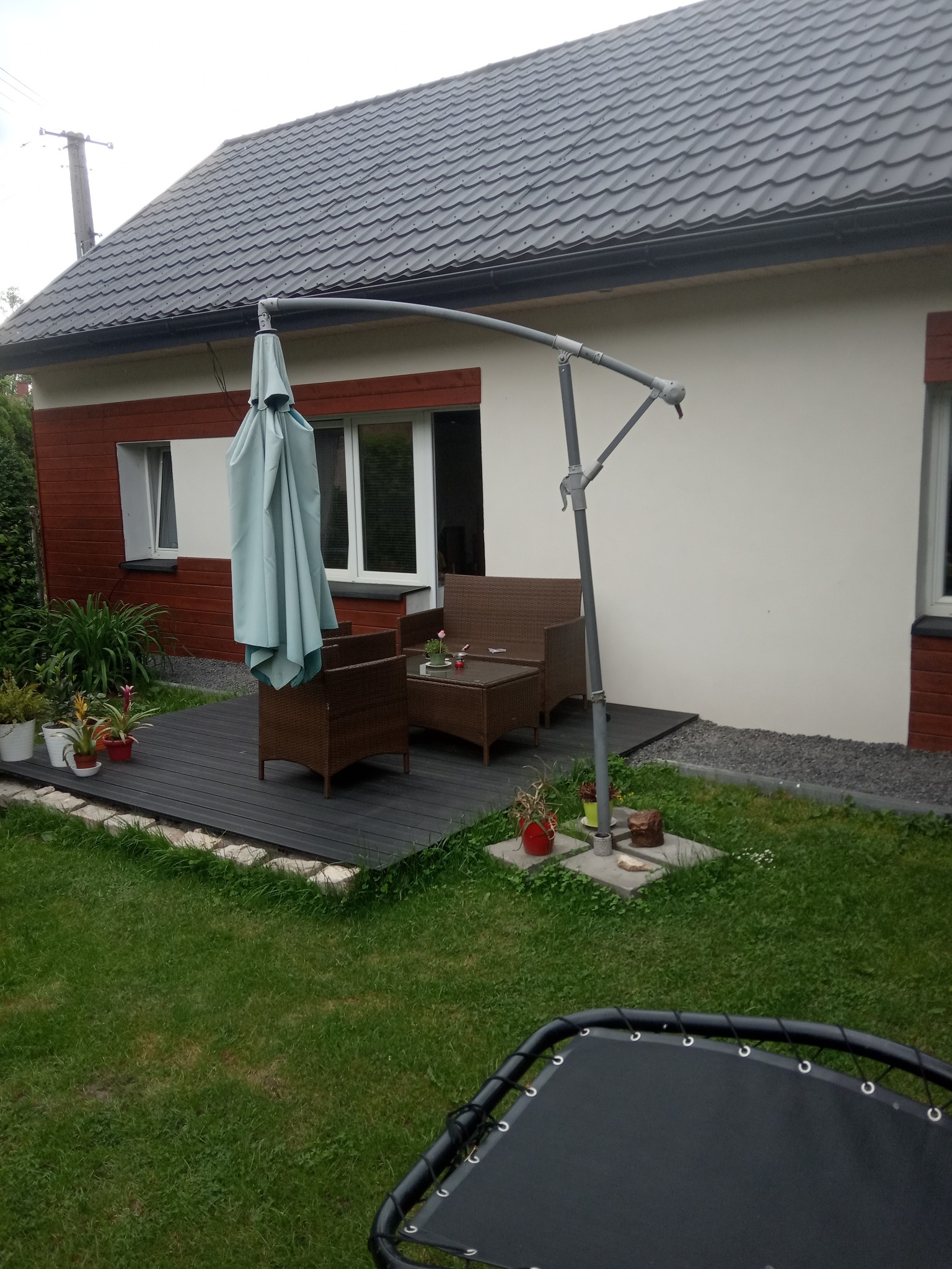 Dom z jasną elewacją i drewnianą okładziną, widoczny z zewnątrz, z tarasem, meblami ogrodowymi i złożonym parasolem.