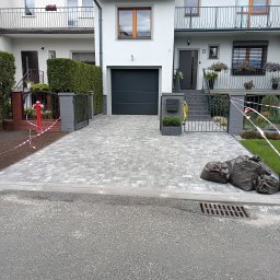 Mariusz Urbanski - Nowa, szara kostka brukowa na podjeździe przed domem z garażem. Widoczne czarne worki z resztkami materiałów budowlanych i taśma ostrzegawcza.