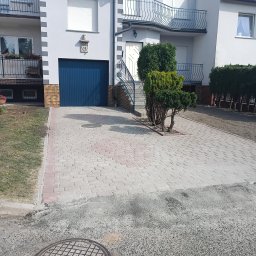 Mariusz Urbanski - Jasnoszara kostka brukowa z czerwoną obwódką na podjeździe przed domem z niebieską bramą garażową i zielonymi krzewami. Widoczny fragment trawnika i studzienka.