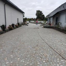 Mariusz Urbanski - Ułożona kostka granitowa na podjeździe, widoczne budynki i pracownik z narzędziami. Całość w stonowanej kolorystyce, z elementami zieleni i szarym niebem.