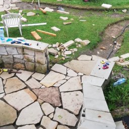 Mariusz Urbanski - Budowa kamiennego tarasu w ogrodzie: nieregularne płyty kamienne i stopnie z jasnego kamienia, narzędzia pracy, taczka i zabawki dziecięce na trawie w tle.