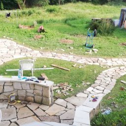 Mariusz Urbanski - Budowa tarasu kamiennego w ogrodzie: widoczny fragment tarasu z jasnych, nieregularnych kamieni, narzędzia pracy, białe krzesło i wózek transportowy na trawniku, w tle las.