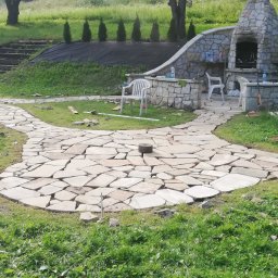 Mariusz Urbanski - Nieregularny kamienny chodnik w trakcie budowy na trawniku, z widocznym ogniskiem z kamienia, narzędziami i białymi plastikowymi krzesłami w tle.