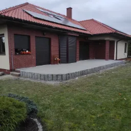 Wykończony taras z kamienną obudową przy domu z panelami słonecznymi na dachu, widok na trawnik i krzewy.