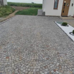 Ułożona kostka granitowa w kształcie wachlarza na podjeździe przed domem z widocznym trawnikiem w tle i małym psem przy drzwiach wejściowych.