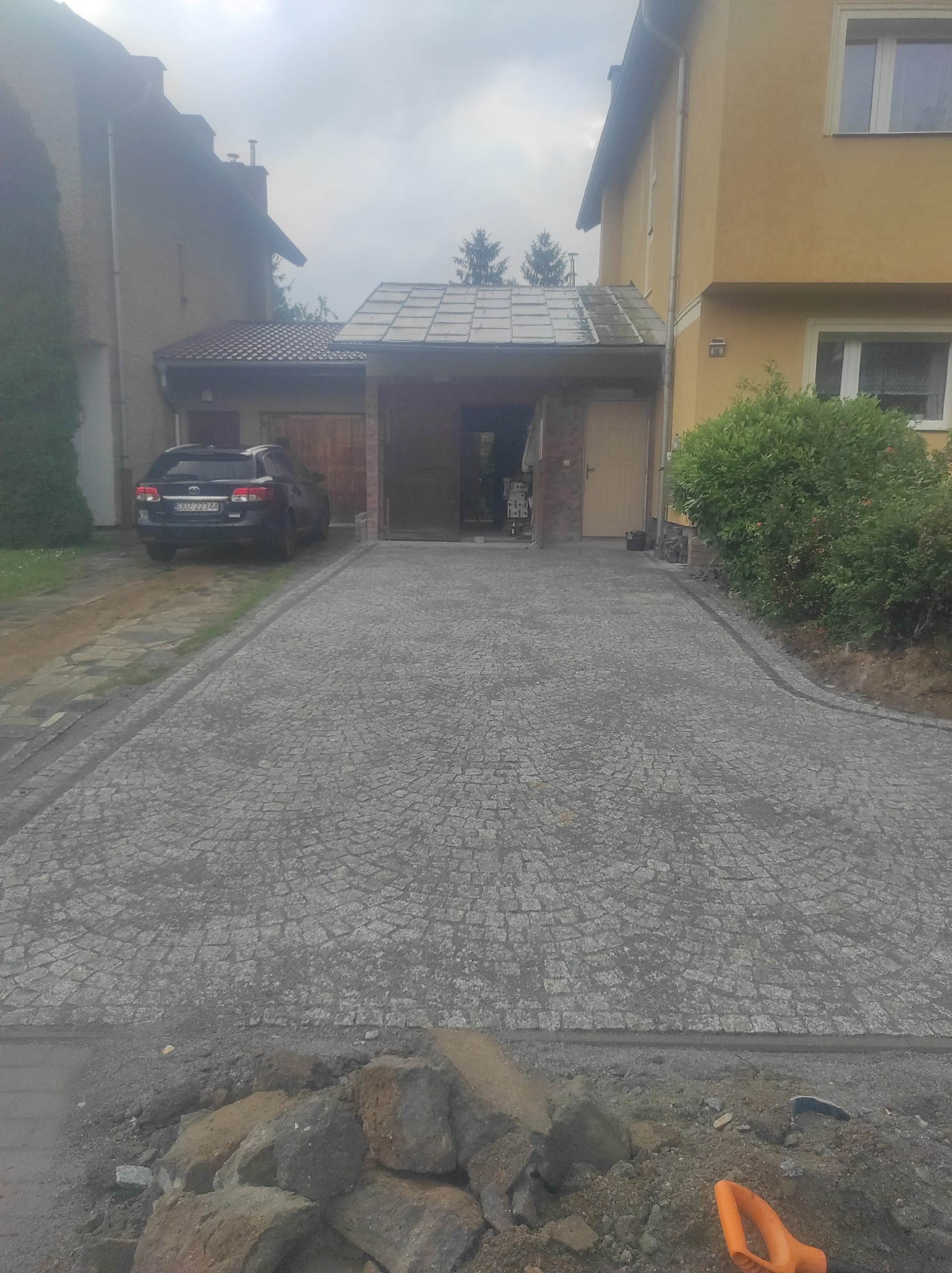 Ułożona szara kostka granitowa tworząca podjazd do garażu między dwoma domami. W tle zaparkowany samochód. Na pierwszym planie narzędzia brukarskie.