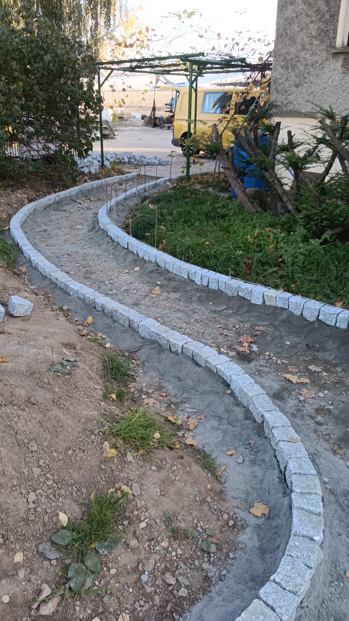 Wąska, kręta ścieżka z jasnoszarej kostki granitowej w trakcie budowy, z widocznymi elementami podbudowy i luźno ułożonymi kostkami obok. Tło stanowi ogród.