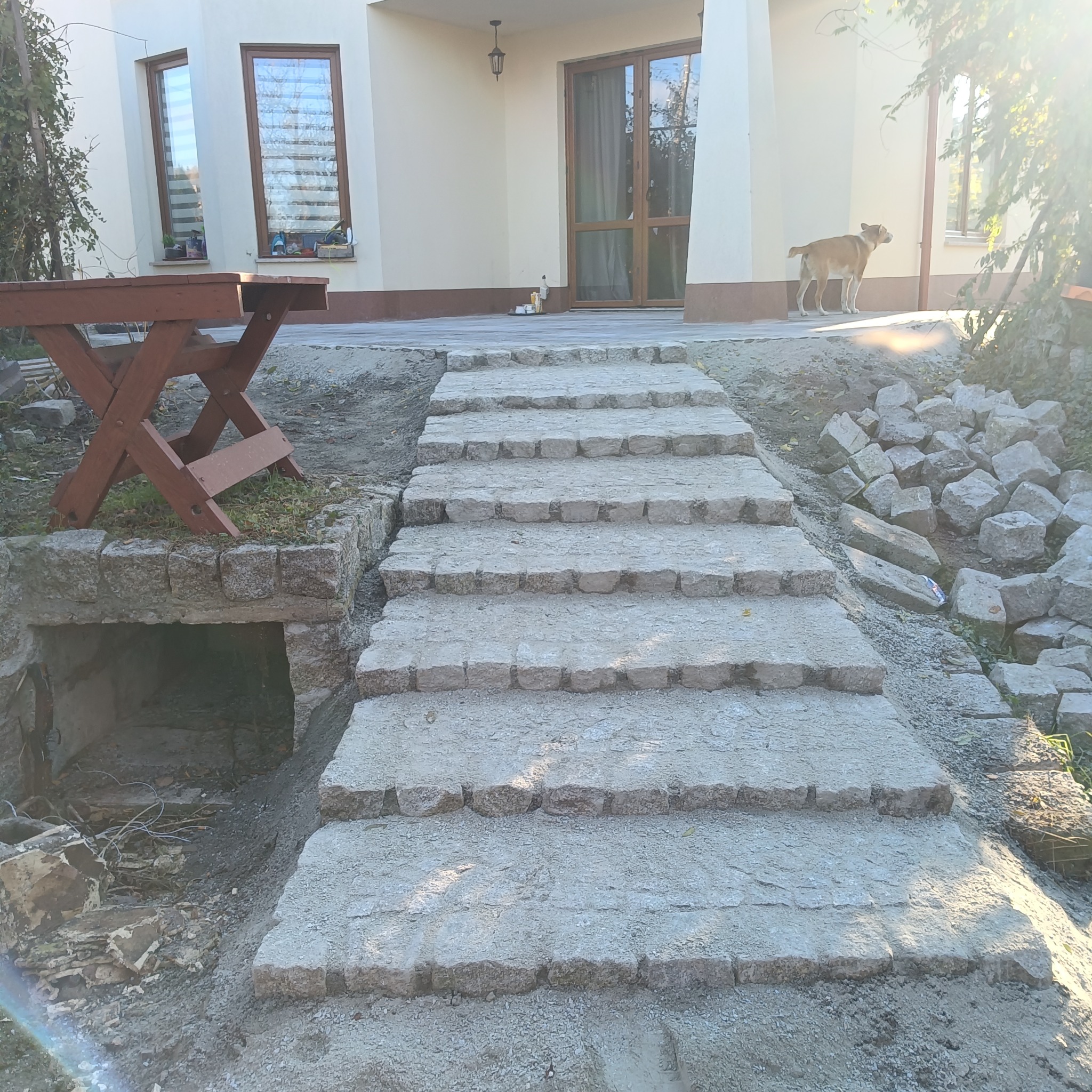 Kamienne schody prowadzące do domu, z widocznym fragmentem tarasu i stertą kostki brukowej obok. Pies stoi na tarasie. Widoczne okna i drzwi domu.