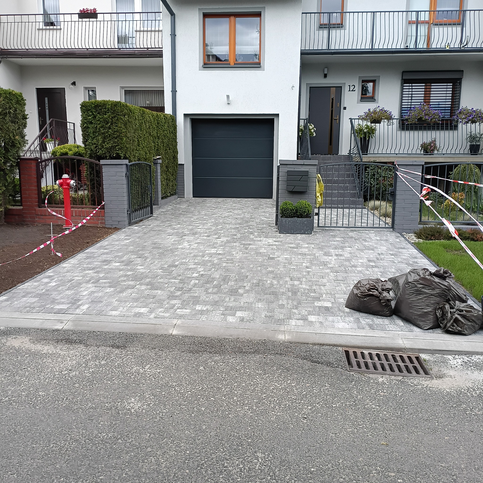 Nowa, szara kostka brukowa na podjeździe przed domem z garażem. Widoczne czarne worki z resztkami materiałów budowlanych i taśma ostrzegawcza.