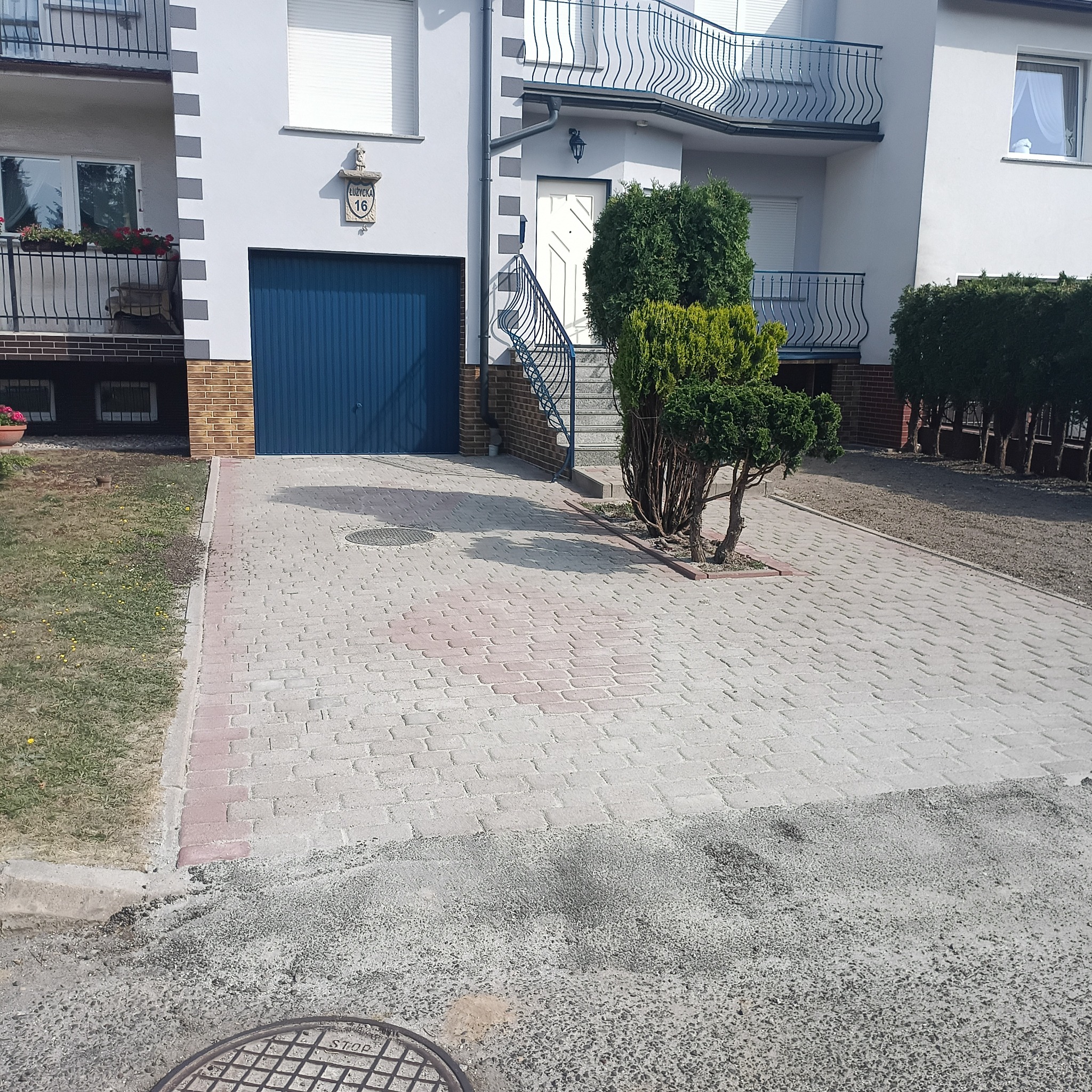 Jasnoszara kostka brukowa z czerwoną obwódką na podjeździe przed domem z niebieską bramą garażową i zielonymi krzewami. Widoczny fragment trawnika i studzienka.