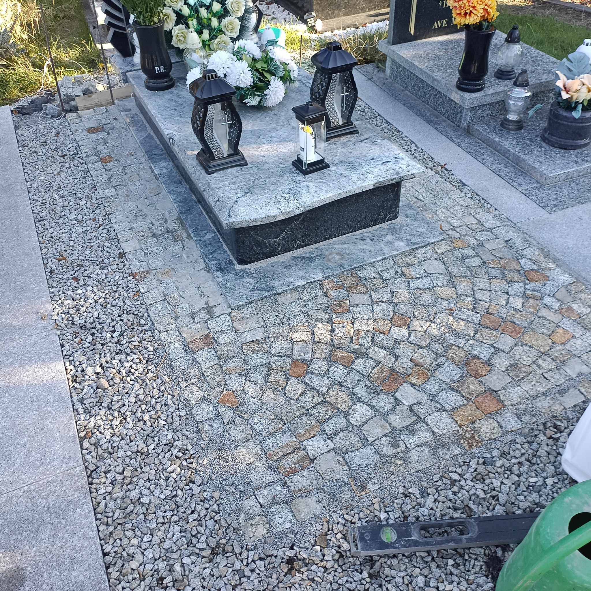 Granitowa kostka ułożona na cmentarzu, tworząca wzór wachlarza. Widoczne nagrobki, lampiony i kwiaty. Całość utrzymana w szarościach, z elementami brązu w kostce.