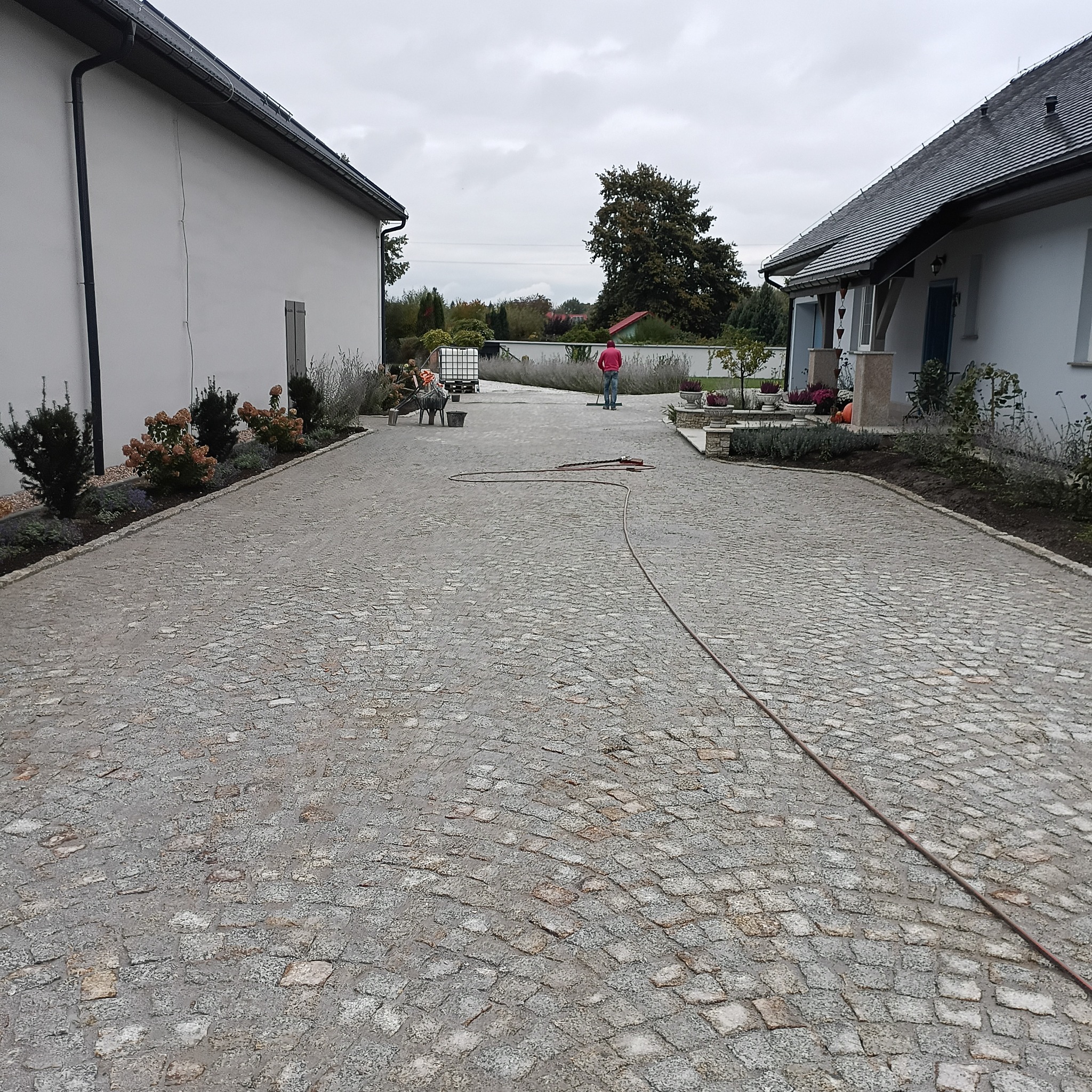 Ułożona kostka granitowa na podjeździe, widoczne budynki i pracownik z narzędziami. Całość w stonowanej kolorystyce, z elementami zieleni i szarym niebem.