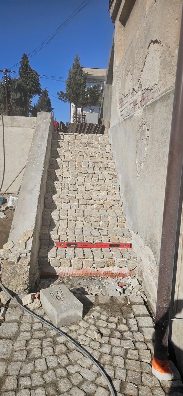 Budowa schodów z kostki granitowej. Widoczny poziomica, fragment chodnika z kostki i ściana budynku. Etap prac w jasnym świetle dziennym.