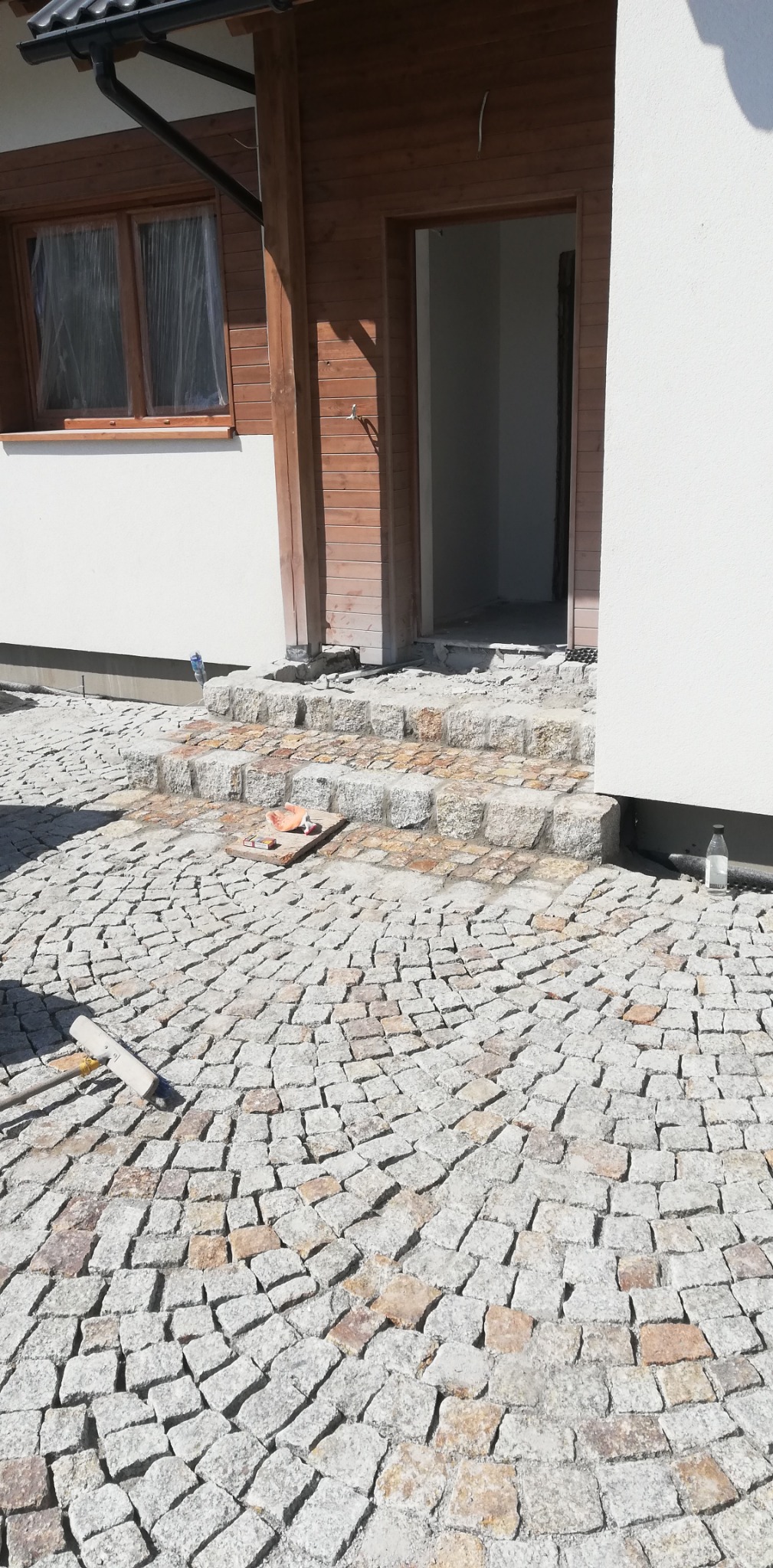 Ułożona kostka granitowa w kształcie wachlarza przed domem, widoczne trzy granitowe stopnie wejściowe, na stopniu leżą resztki zaprawy i fragmenty kostki, obok butelka wody i miotła.