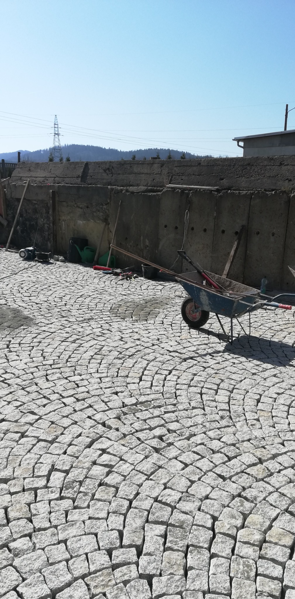 Ułożona z jasnoszarej kostki granitowej nawierzchnia w kształcie wachlarza, z widocznym betonowym murem oporowym w tle i niebieską taczką wypełnioną materiałem budowlanym oraz narzędziami.