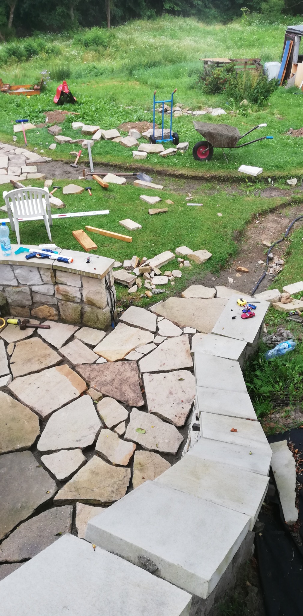 Budowa kamiennego tarasu w ogrodzie: nieregularne płyty kamienne i stopnie z jasnego kamienia, narzędzia pracy, taczka i zabawki dziecięce na trawie w tle.