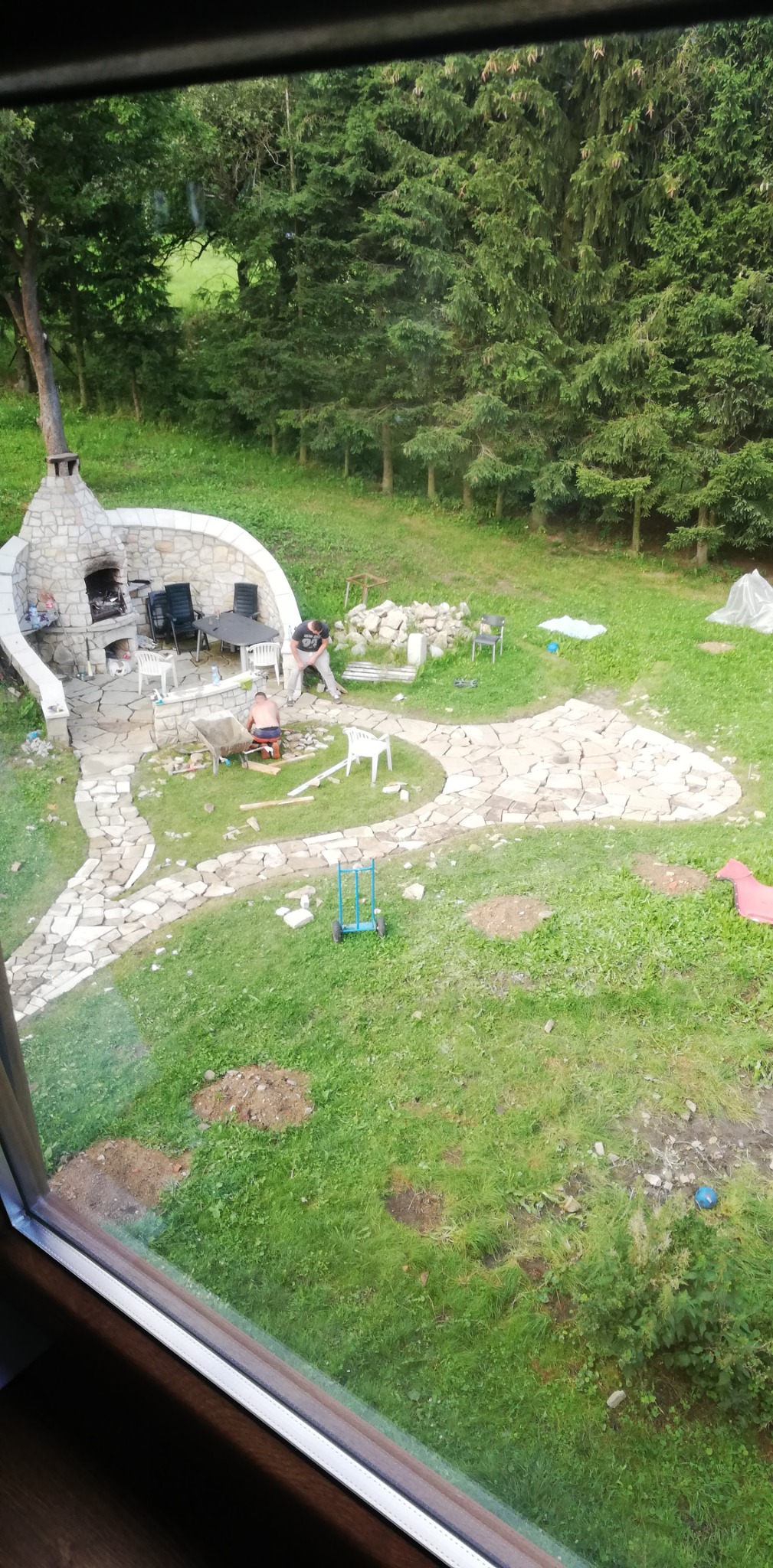 Budowa kamiennego tarasu i ścieżki w ogrodzie z widocznym paleniskiem i dwoma pracownikami układającymi kamienie