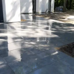 Świeżo ułożona, mokra, szara nawierzchnia z płytek betonowych, odbijająca światło słoneczne i korony drzew, sąsiadująca z ziemią i elementami instalacji.