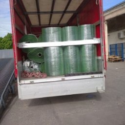 S-trans - Wnętrze ciężarówki z czerwonym plandekowym dachem, załadowane rolkami sztucznej trawy zabezpieczonymi folią stretch na metalowych półkach.