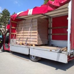 S-trans - Czerwony samochód dostawczy z otwartymi drzwiami kabiny, załadowany paletą z drewnianymi elementami zabezpieczonymi pasami, widoczne czerwone wnętrze skrzyni ładunkowej i częściowo zwinięta plandeka.