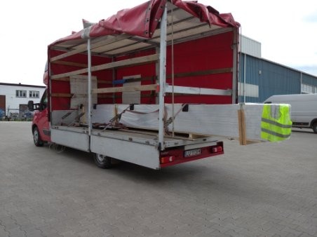 Czerwony samochód dostawczy z otwartą przestrzenią ładunkową przewozi paletę białych płyt, zabezpieczonych pasami, z widoczną kamizelką odblaskową na ładunku.