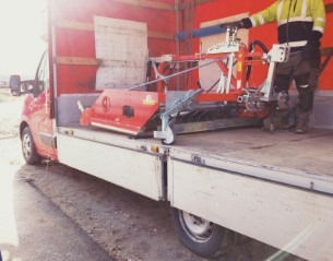 Czerwony, specjalistyczny sprzęt rolniczy zabezpieczony pasami w przestrzeni ładunkowej samochodu dostawczego z otwartą burtą. Widoczny fragment sylwetki pracownika w odblaskowej kamizelce.