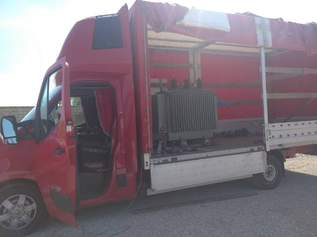 Czerwony samochód dostawczy z otwartą przestrzenią ładunkową, w której znajduje się duży transformator elektryczny. Widoczne czerwone zasłony w kabinie kierowcy.