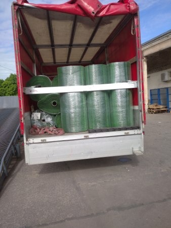 Wnętrze ciężarówki z czerwonym plandekowym dachem, załadowane rolkami sztucznej trawy zabezpieczonymi folią stretch na metalowych półkach.