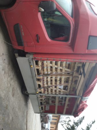 Czerwony samochód dostawczy z otwartą przestrzenią ładunkową wypełnioną drewnianymi elementami konstrukcyjnymi, widoczne koła i fragment kabiny.