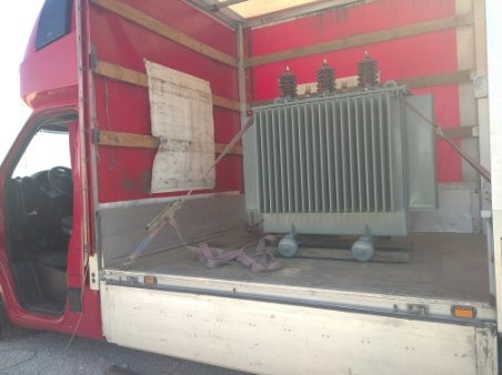 Duży transformator elektryczny zabezpieczony pasami wewnątrz czerwonej furgonetki dostawczej z otwartymi tylnymi drzwiami.