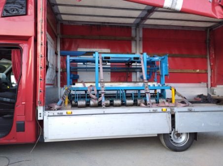 Niebieska maszyna przemysłowa zabezpieczona pasami transportowymi wewnątrz czerwonej ciężarówki z otwartymi drzwiami kabiny.