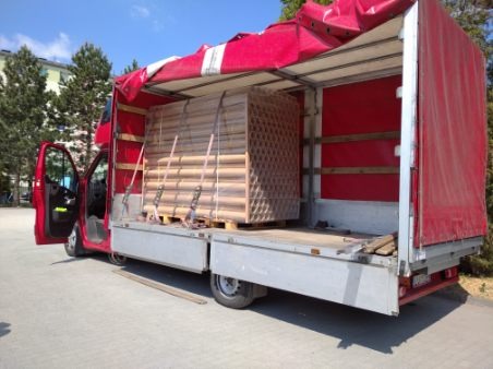 Czerwony samochód dostawczy z otwartymi drzwiami kabiny, załadowany paletą z drewnianymi elementami zabezpieczonymi pasami, widoczne czerwone wnętrze skrzyni ładunkowej i częściowo zwinięta plandeka.